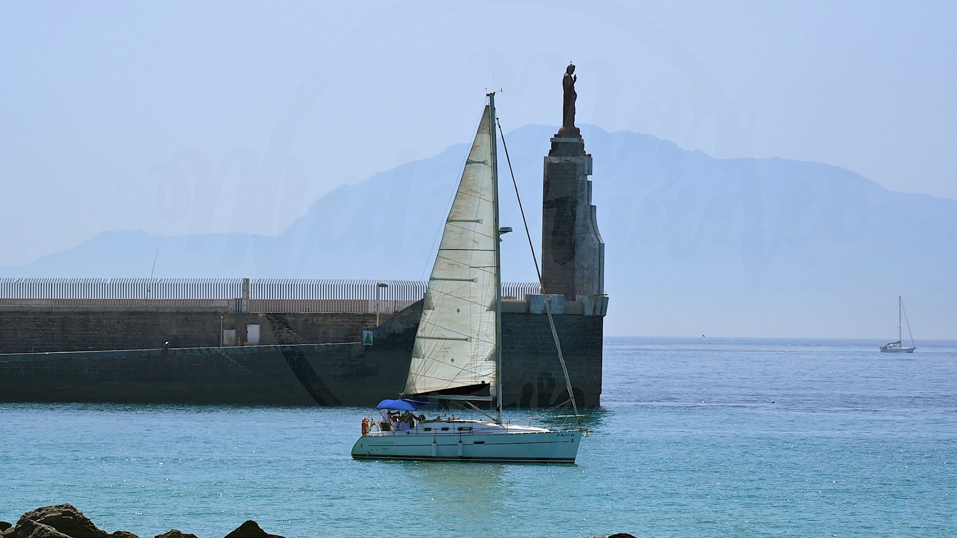 Velero pasando junto al monumento del Sagrado Corazón a la salida del puerto de Tarifa, con las montañas de Marruecos al fondo