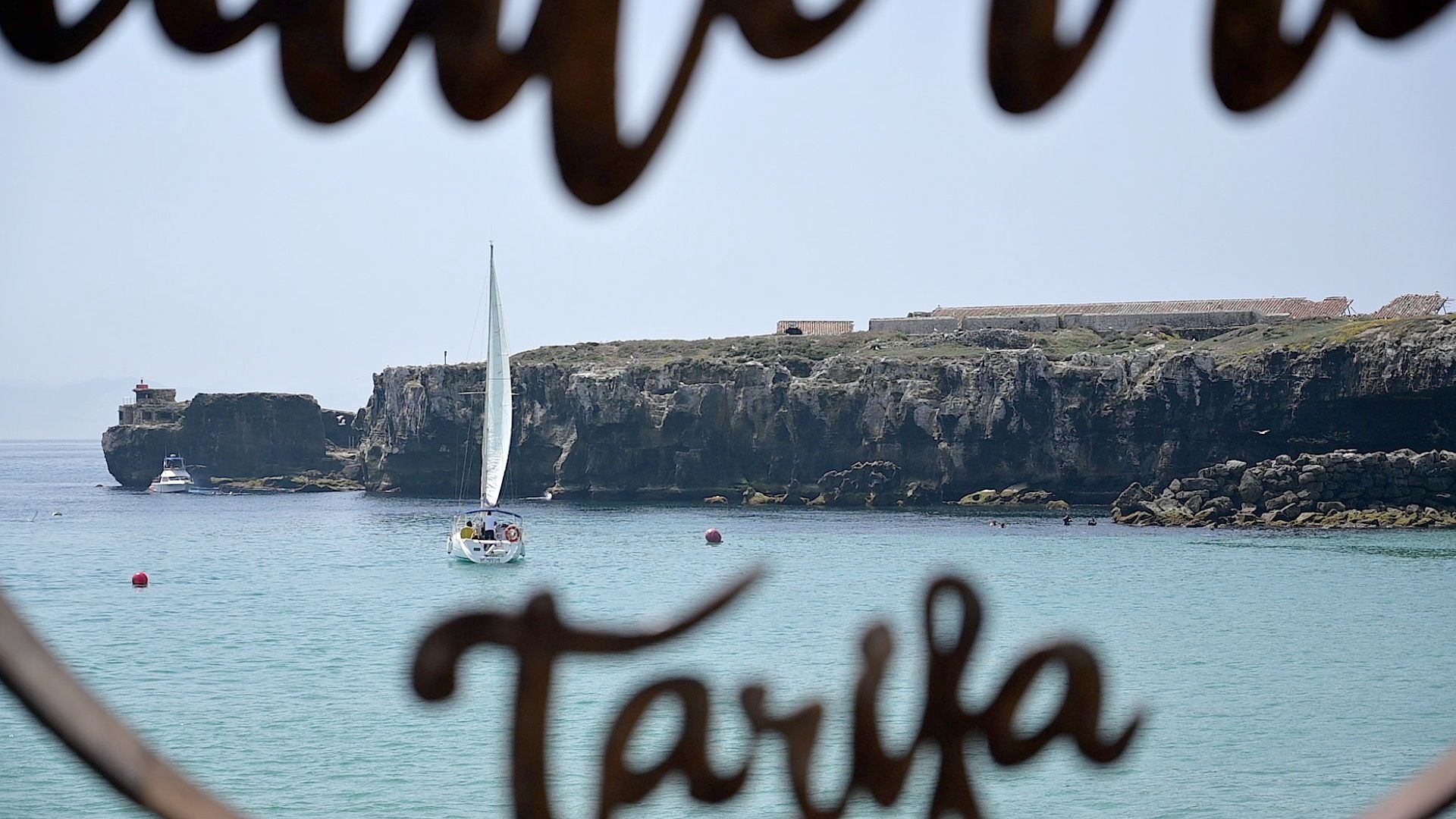 Velero navegando junto a los acantilados de la Isla de las Palomas, visto a través de la escultura de Tarifa