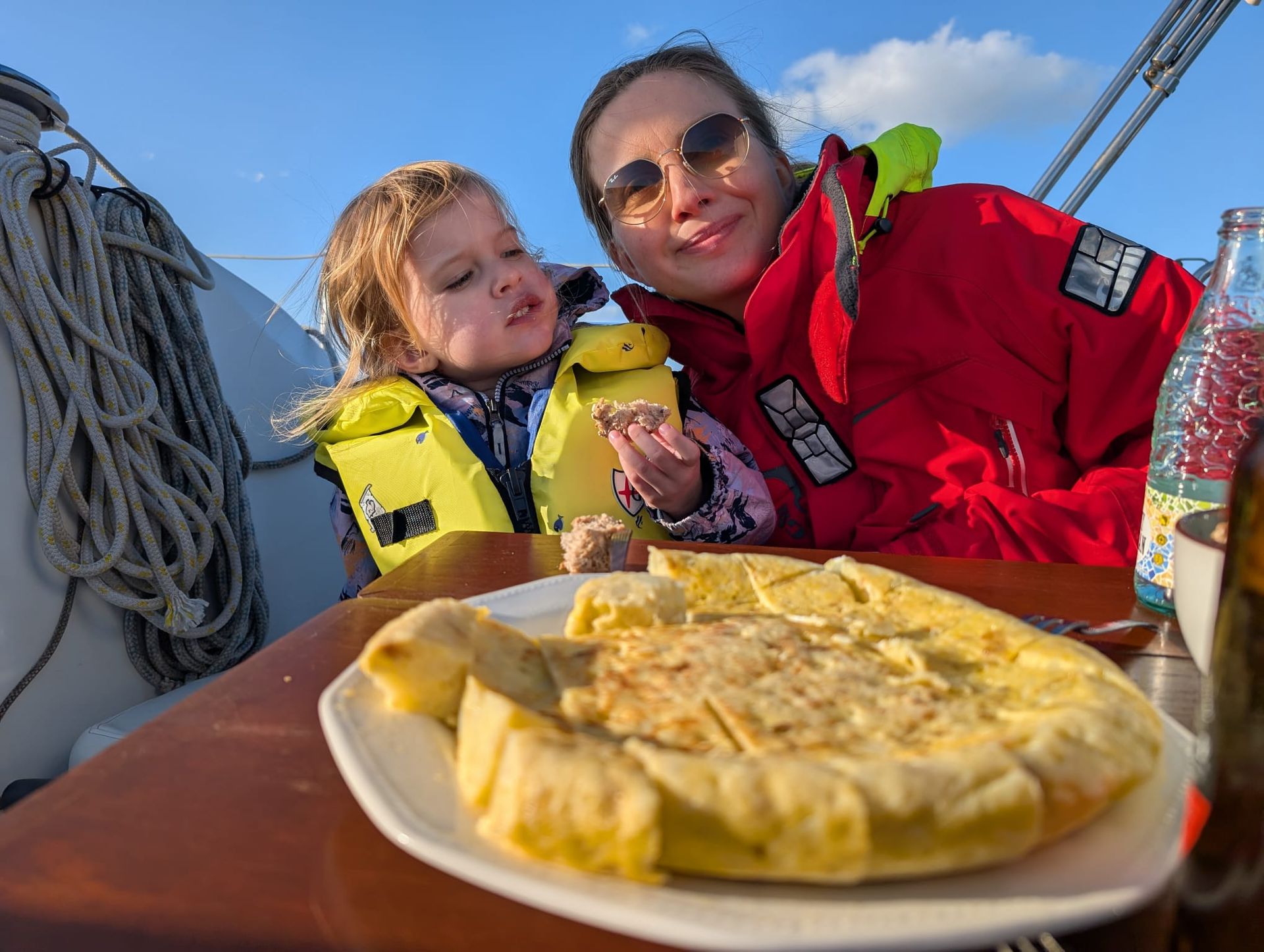 A mother and her toddler in lifejackets sharing homemade tortilla on board under a blue sky