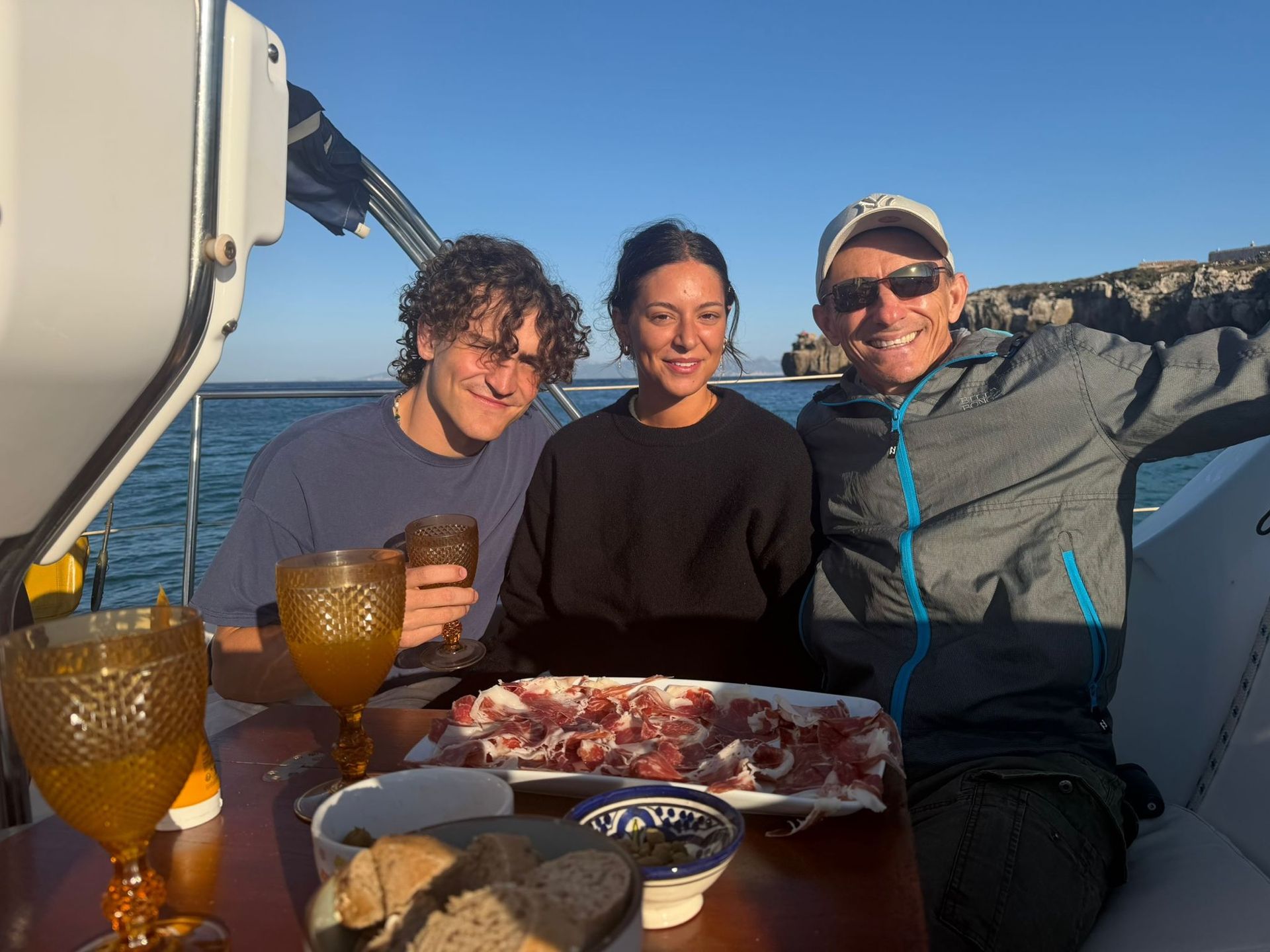 Three guests enjoying jamón and tapas with golden glasses at sunset, the cliffs of Tarifa behind