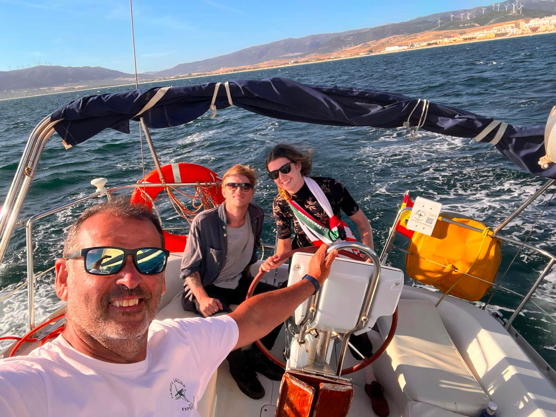 El patrón y dos pasajeros al timón navegando frente a los molinos de viento de Tarifa